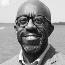 Headshot of a Black man with glasses smiling and standing in front of a body of water